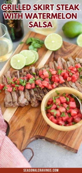 A grilled skirt steak with watermelon salsa is sliced on a wooden board, topped with the vibrant salsa. Additional watermelon salsa in a bowl sits beside, accompanied by lime wedges and cilantro in the background.