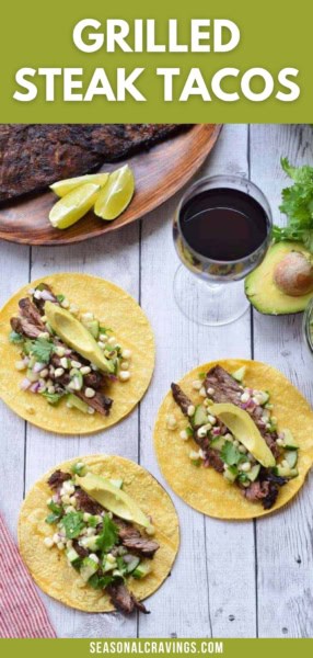 Three grilled steak tacos topped with corn, avocado slices, cilantro, and onion on a wooden surface, with a glass of red wine, lime wedges, an avocado, and a grilled steak in the background.