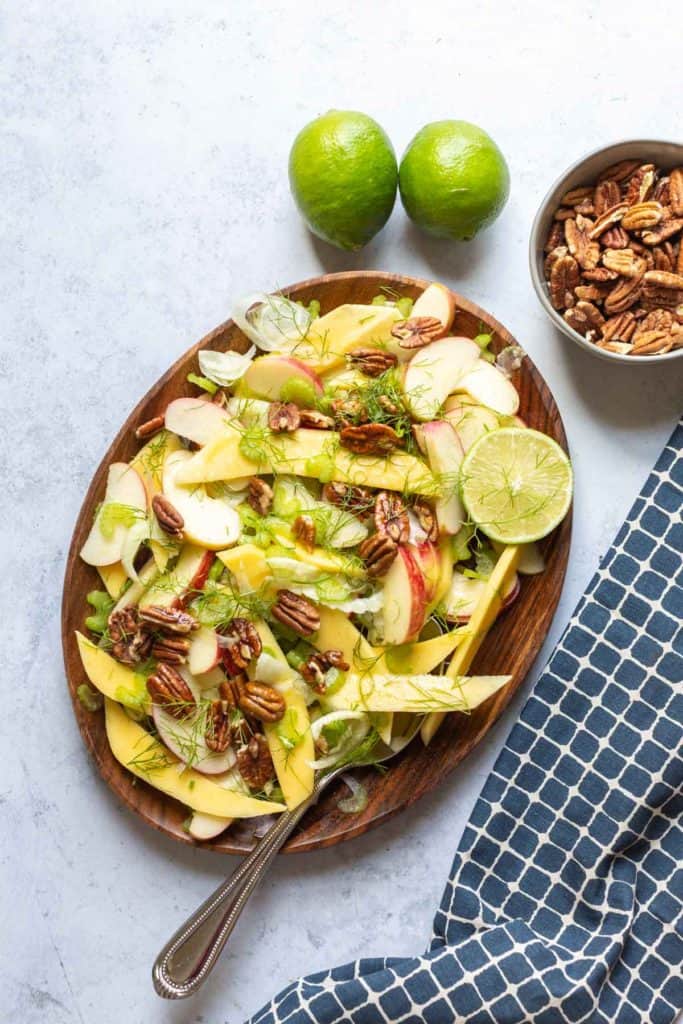 mango apple and fennel salad on a brown plate with bowl of pecans on side