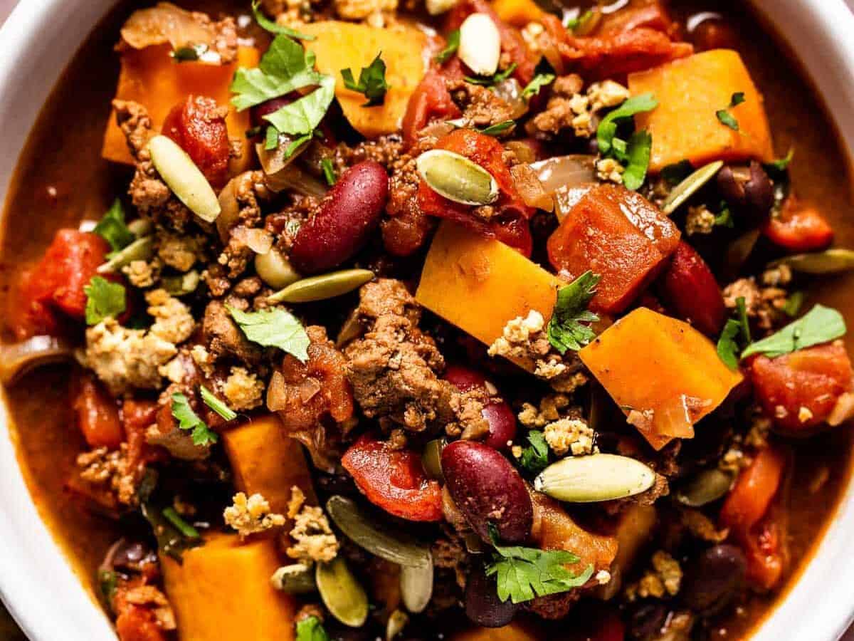 A hearty bowl of chili with ground meat, beans, diced tomatoes, cubed squash, and sprinkled with pumpkin seeds and chopped cilantro.