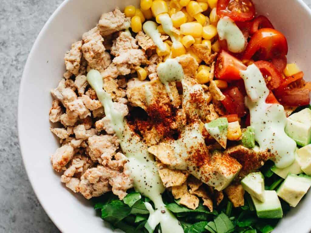 A white bowl containing chopped vegetables, cooked ground meat, corn, diced tomatoes, avocado, greens, and tortilla chips, topped with a green dressing and sprinkled with paprika.