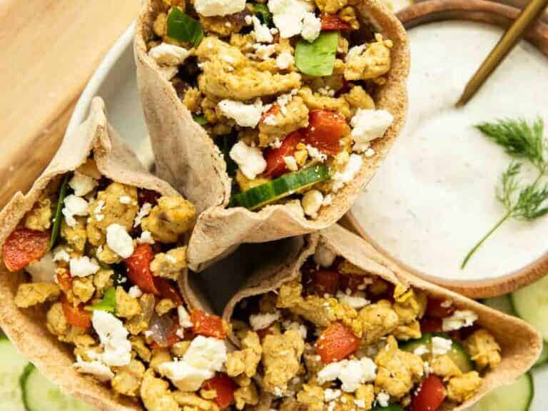 Three pita wraps filled with scrambled eggs, diced vegetables, and crumbled feta cheese are arranged on a plate next to a bowl of dipping sauce with a sprig of dill.