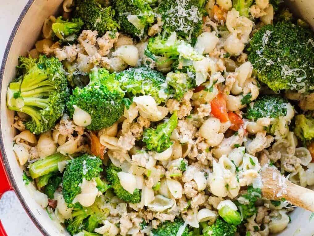 A pot filled with a cooked mixture of pasta shells, broccoli florets, ground meat, and diced tomatoes, all blended together with a wooden spoon.