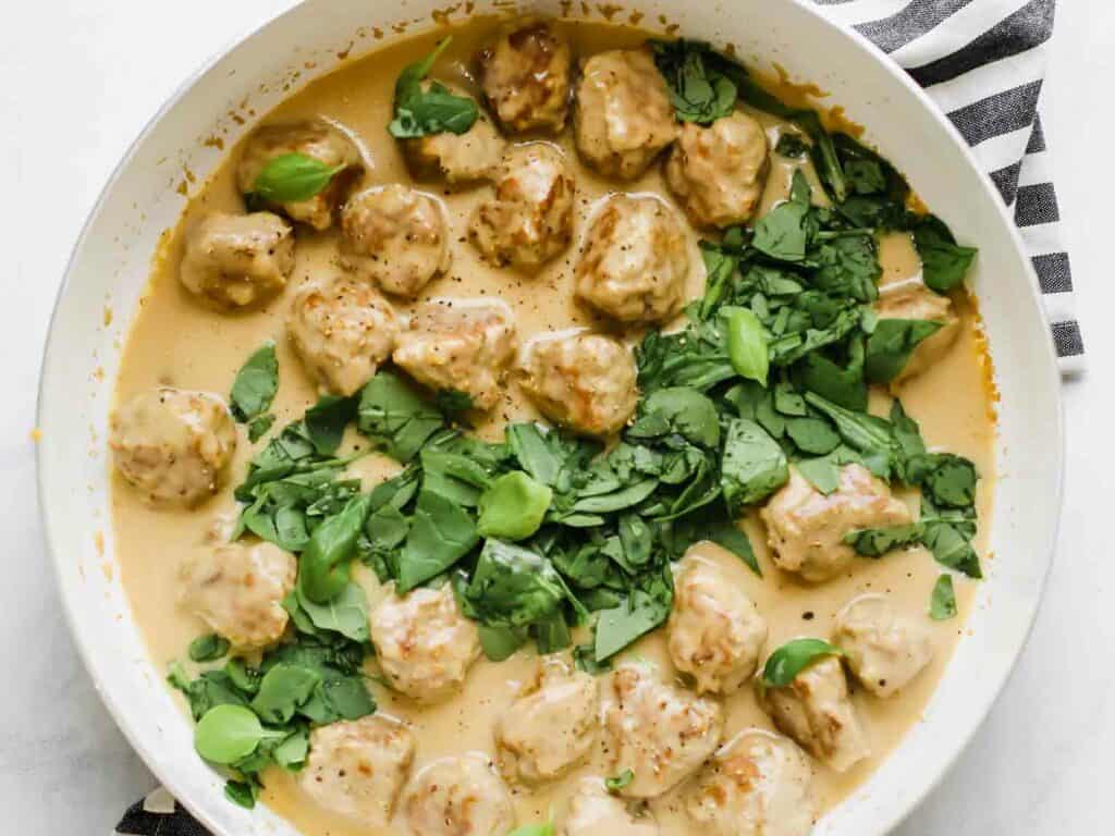 A pan of meatballs in a creamy sauce, topped with chopped green herbs and spinach. The handle of the pan is visible in the corner.