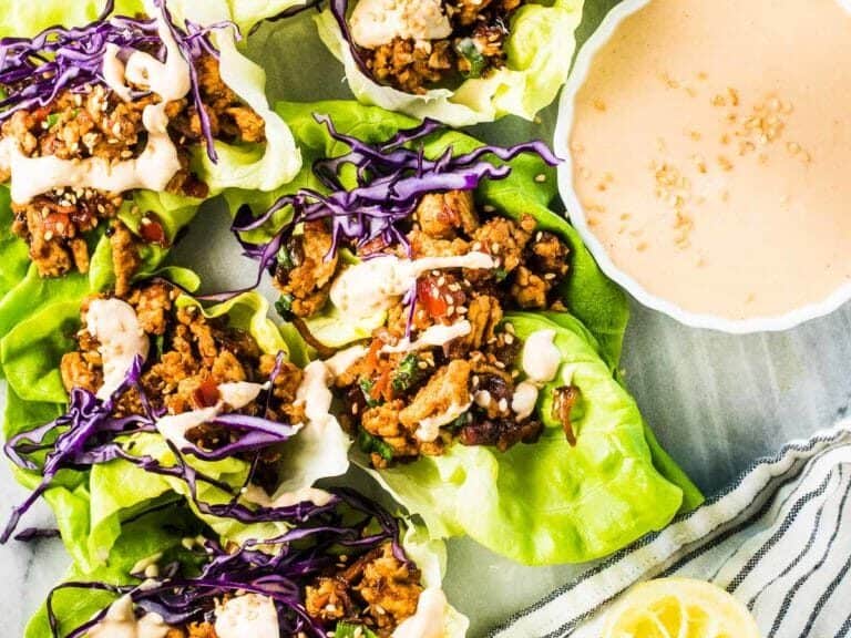 Lettuce wraps filled with seasoned ground meat, shredded purple cabbage, and drizzled sauce next to a bowl of dipping sauce.