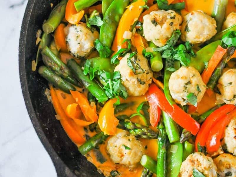 A skillet filled with a colorful vegetable stir-fry featuring snow peas, bell peppers, asparagus, and meatballs in a flavorful sauce.