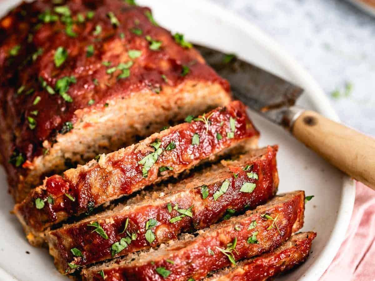 A sliced meatloaf topped with chopped herbs is displayed on a white plate, with a knife placed beside it.