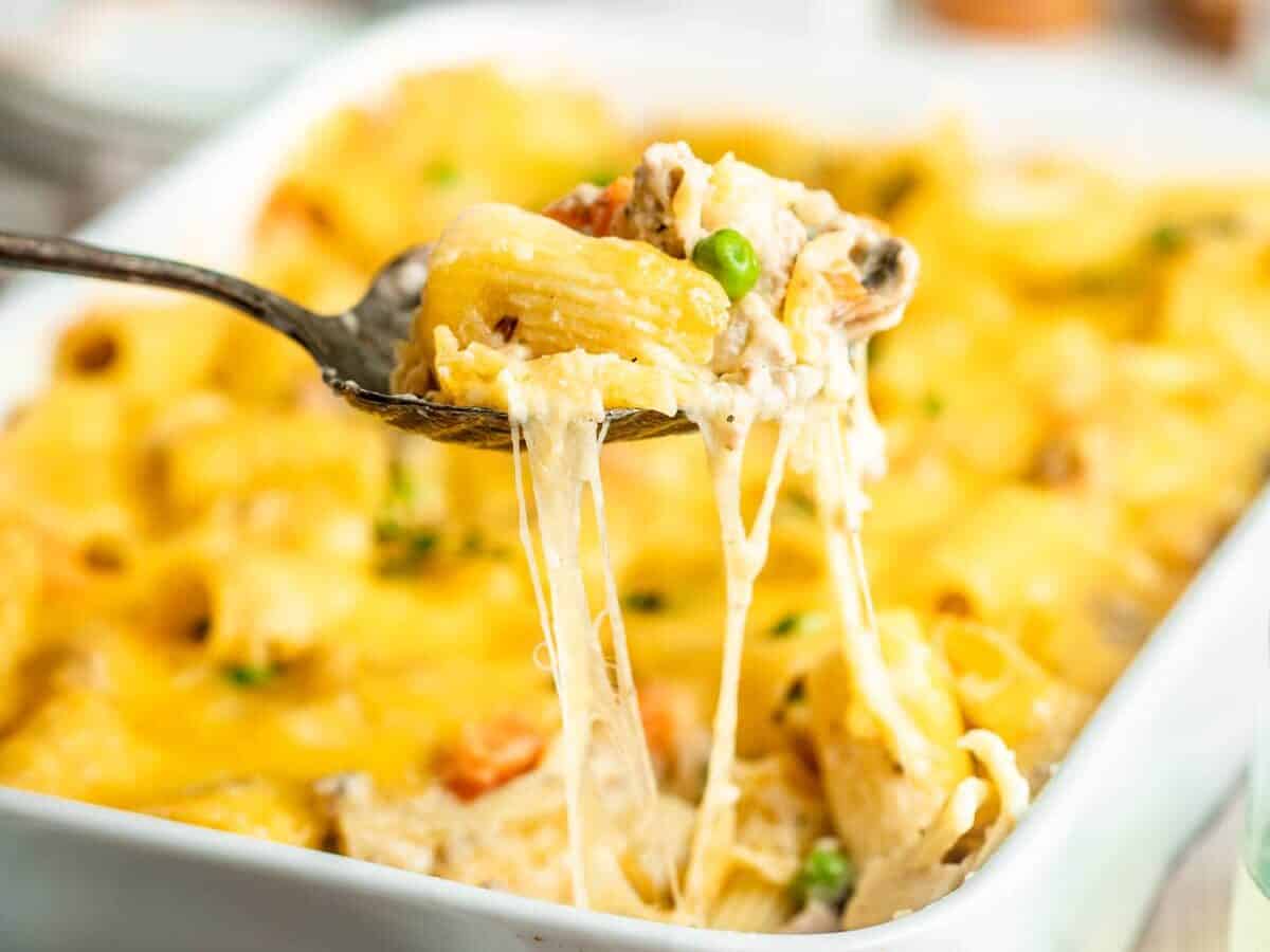 A close-up of a fork lifting a cheesy pasta bake from a casserole dish. The pasta includes vegetables such as peas, carrots, and mushrooms.