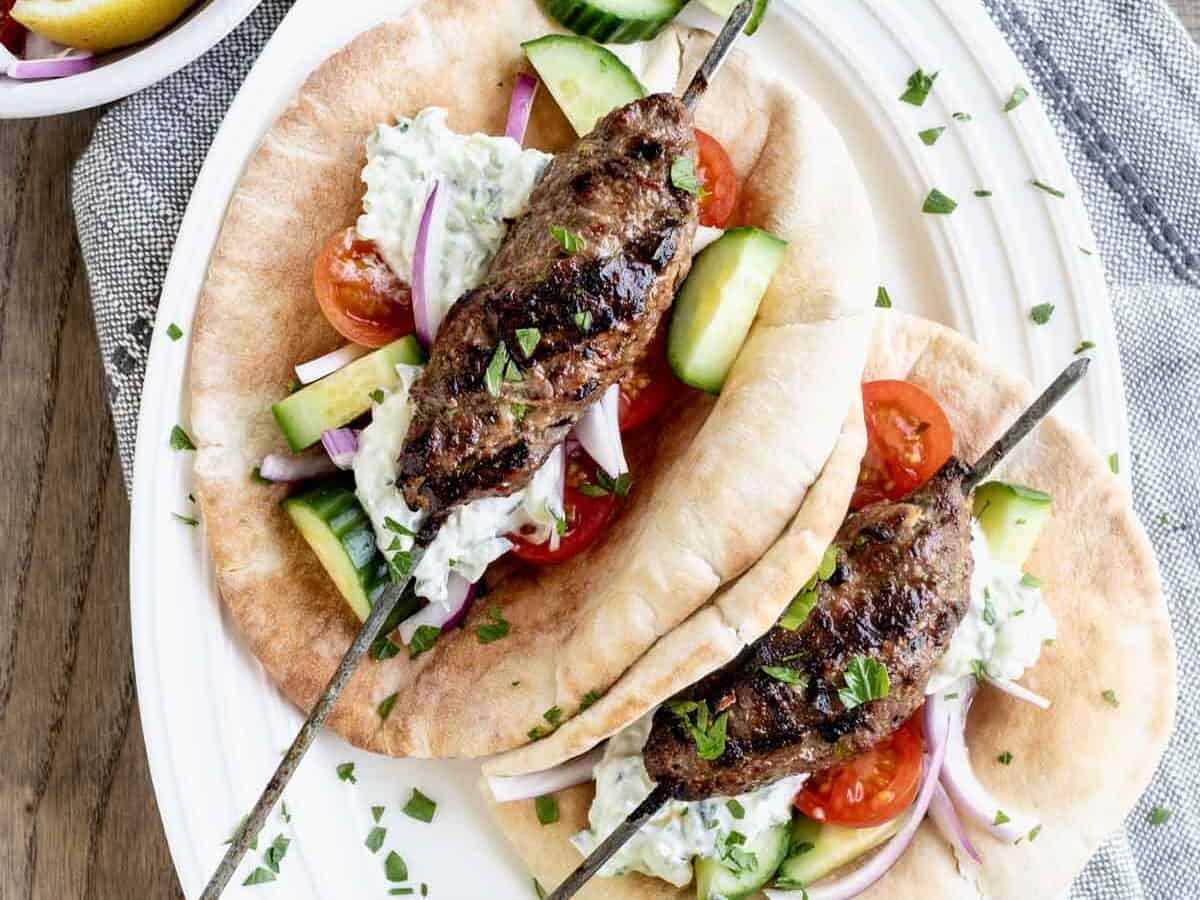 Two pita wraps with grilled meat skewers, tzatziki sauce, sliced cucumbers, tomatoes, and red onions on a white plate.