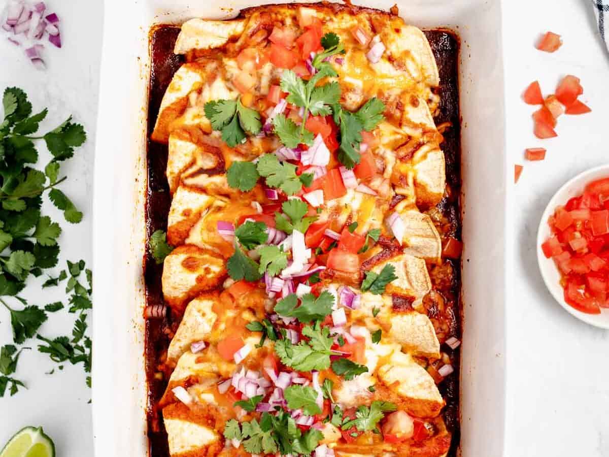 A baking dish filled with enchiladas topped with melted cheese, chopped tomatoes, red onions, and cilantro.