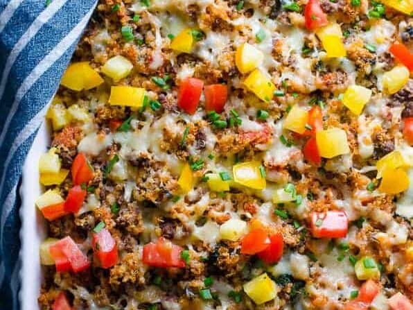 A close-up view of a baked dish topped with melted cheese, colorful diced bell peppers, chopped green onions, and ground meat, with a striped cloth partially visible in the corner.