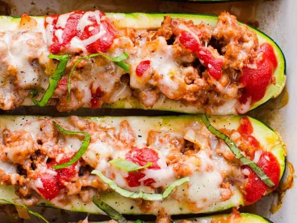 Close-up of zucchini boats stuffed with a ground meat mixture, topped with melted cheese, tomato sauce, and garnished with thinly sliced fresh basil.