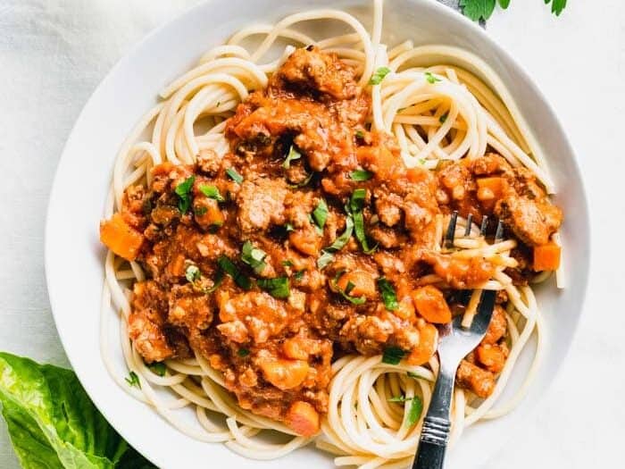 A white bowl filled with spaghetti topped with a chunky tomato and meat sauce, garnished with chopped herbs. A fork is resting in the bowl.