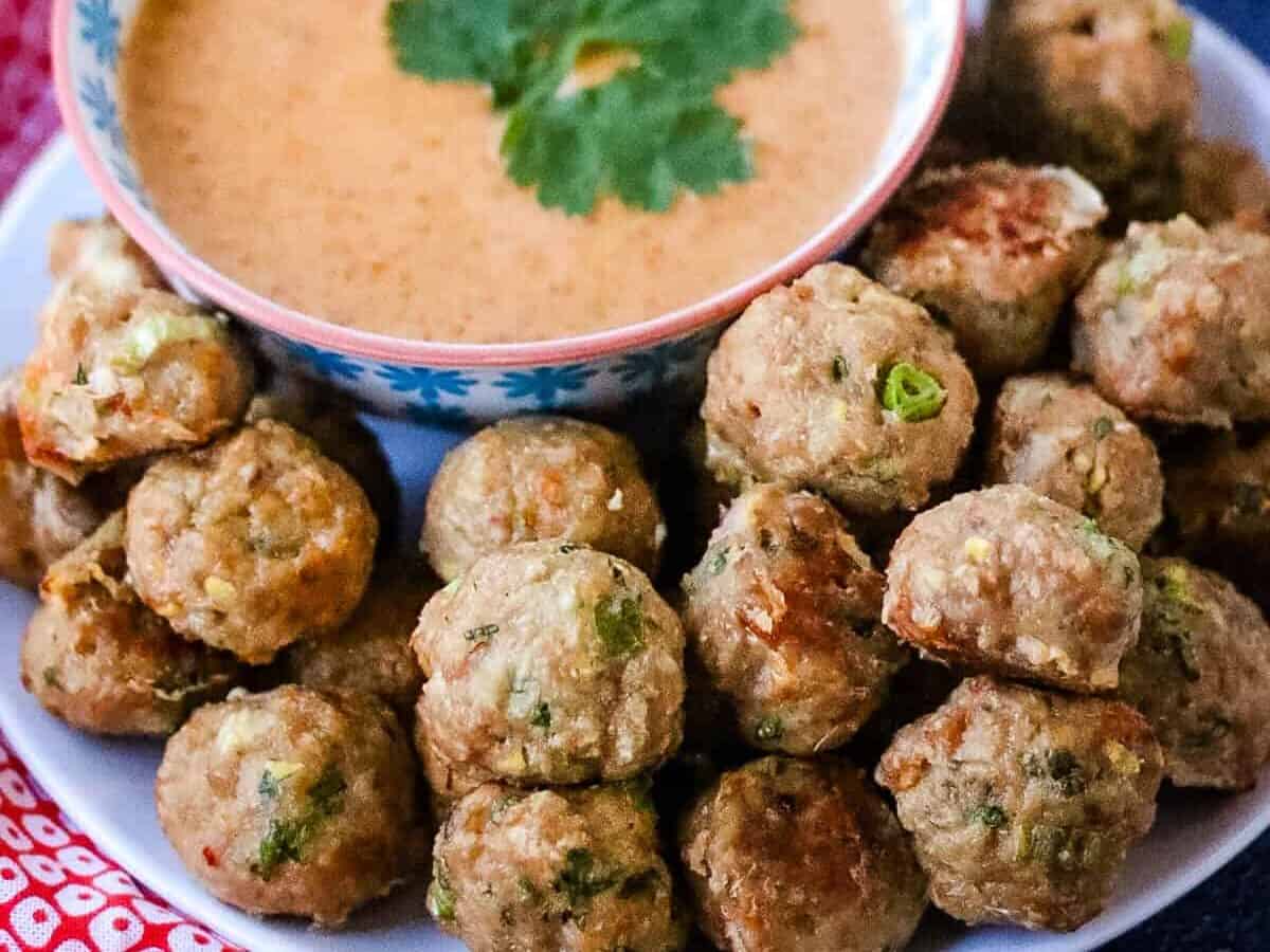 A plate of meatballs with herbs is served around a bowl of dipping sauce garnished with a sprig of cilantro.