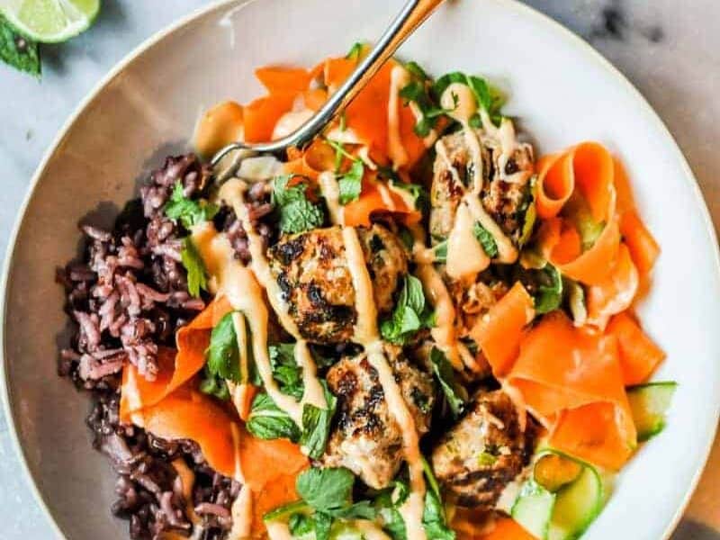 A bowl of black rice, grilled meatballs, and carrot ribbons garnished with fresh herbs and drizzled with a light sauce. A fork is placed in the bowl.