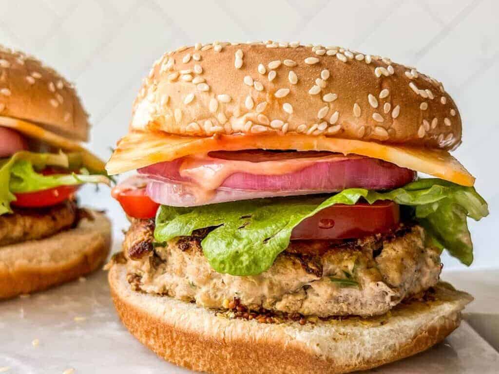 A close-up of a chicken burger with lettuce, tomato, red onion, cheese, and sauce in a sesame seed bun. Another burger is partially visible in the background.