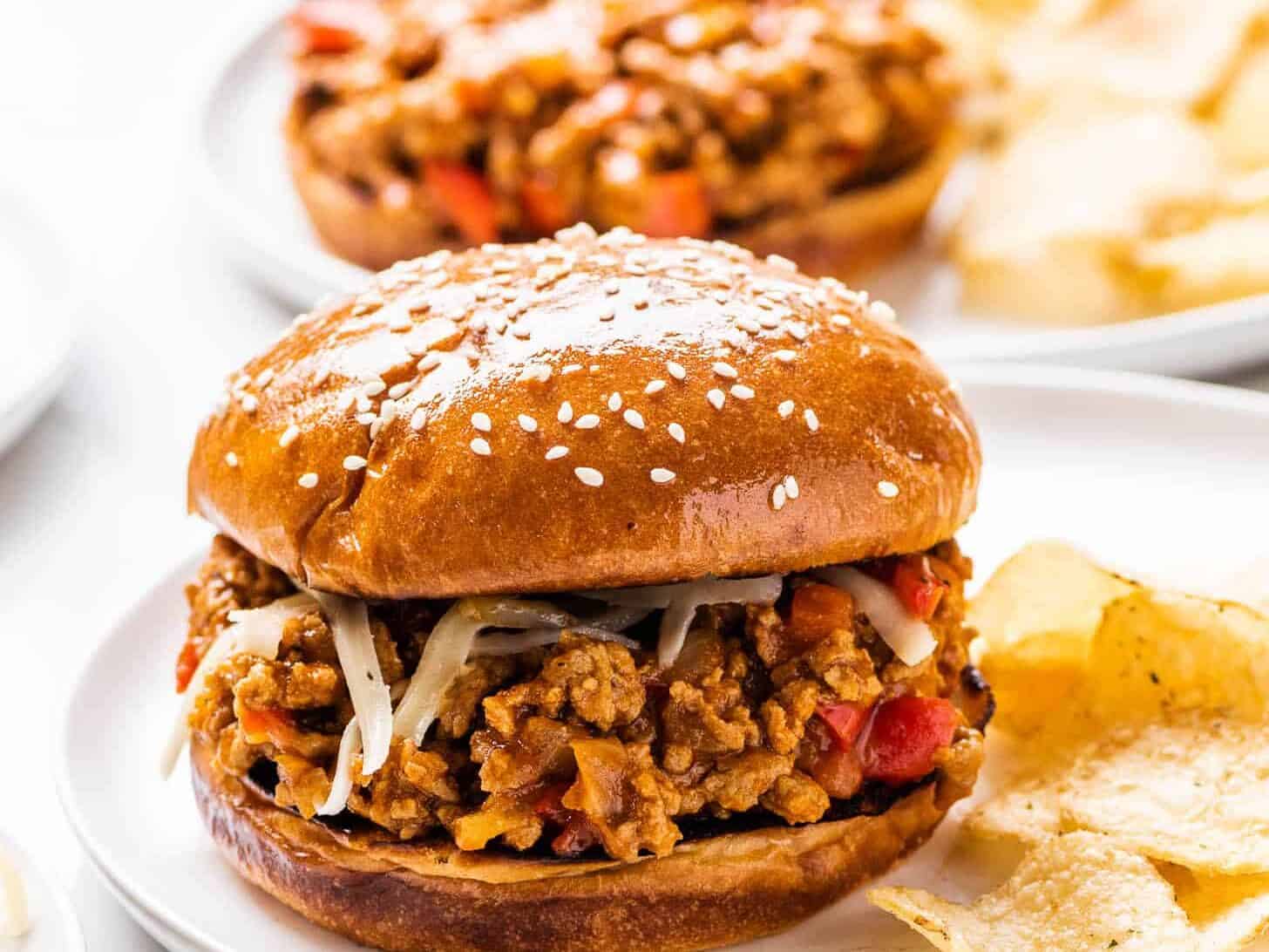 A sesame seed bun sandwich filled with a mixture of ground meat, cheese, and vegetables. Served on a white plate with a side of potato chips.