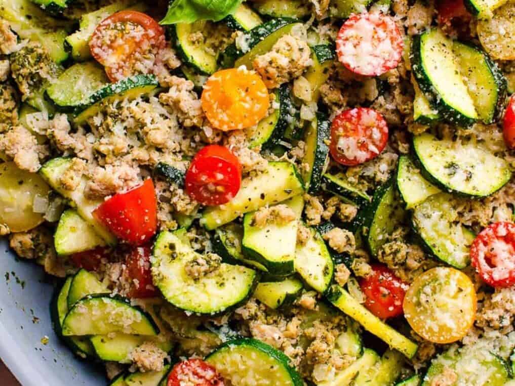 Close-up of a mixed vegetable salad featuring sliced zucchini, cherry tomatoes, and ground meat, garnished with grated cheese and herbs.