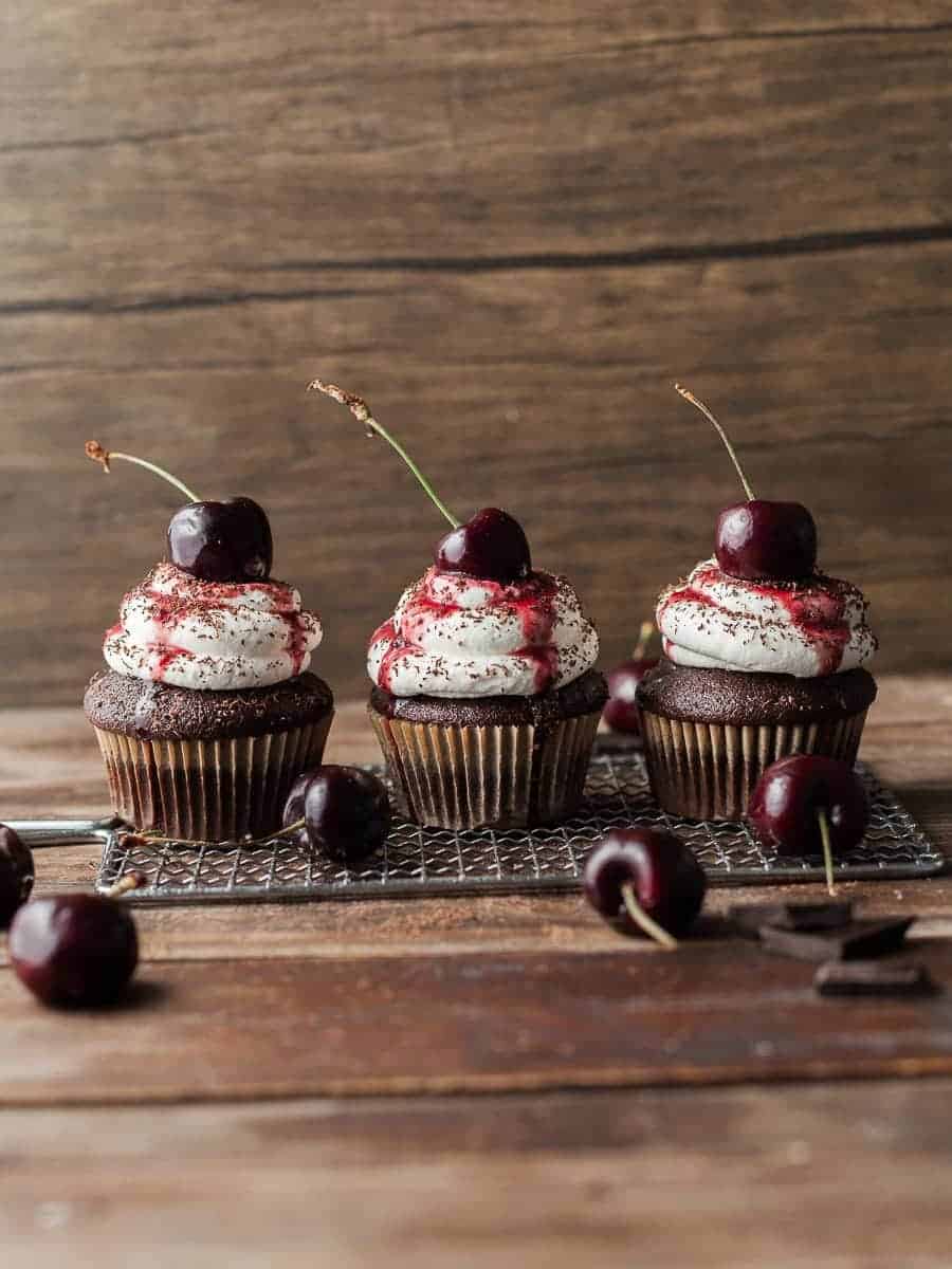 Three decadent chocolate cupcakes topped with a luscious swirl of whipped cream and garnished with juicy cherries on a rustic wooden table.