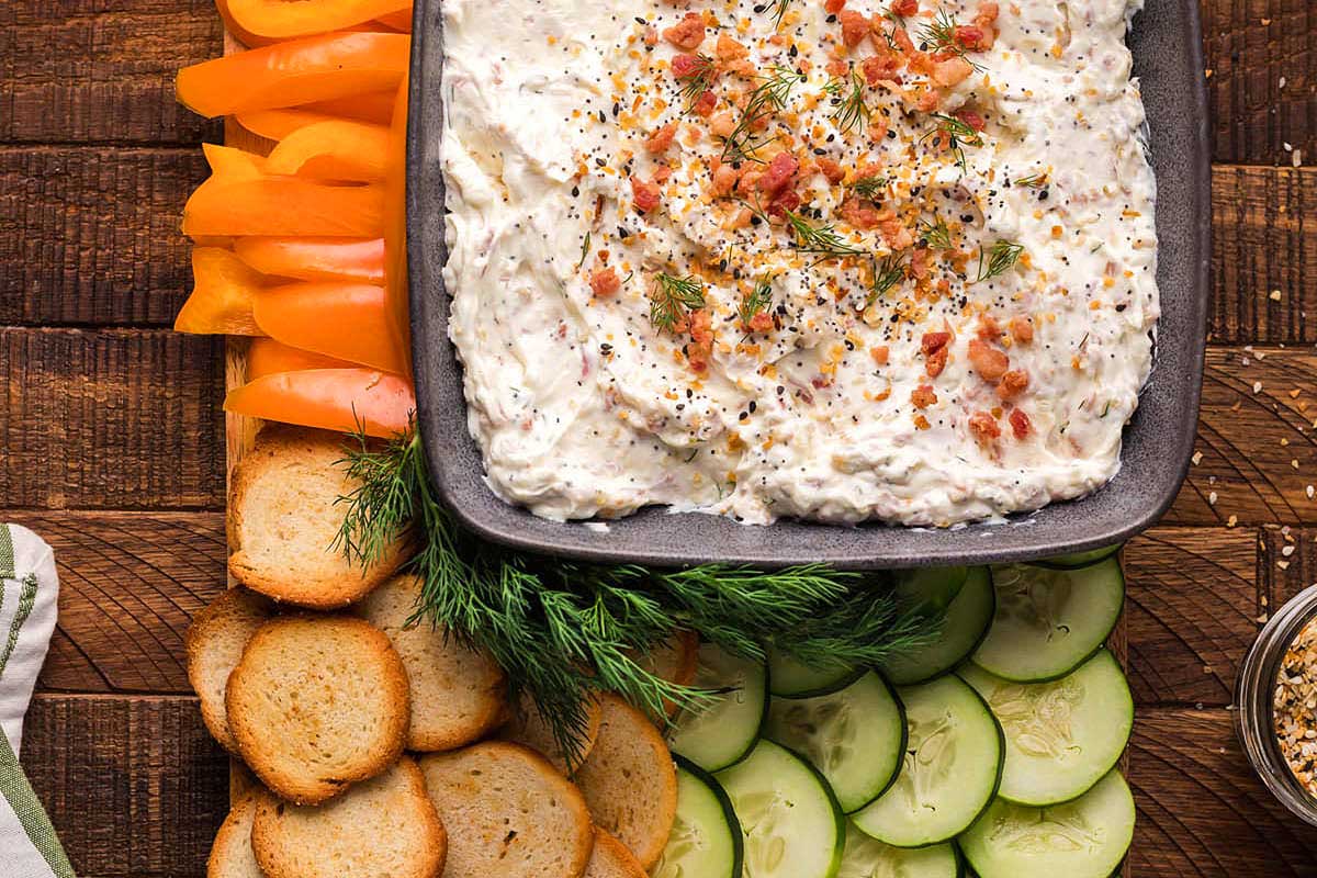 A bowl of dip with vegetables and crackers on a wooden table.