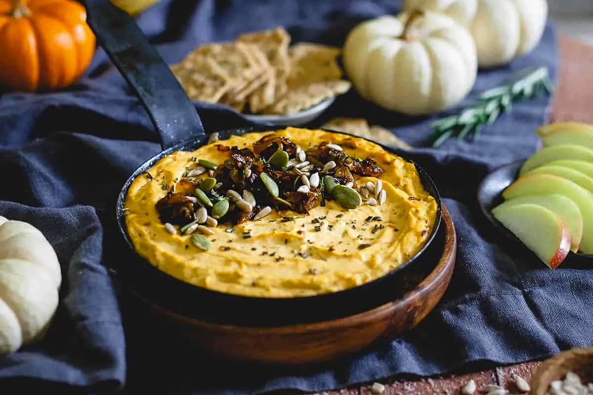 A bowl of pumpkin hummus with pumpkin seeds and apples.