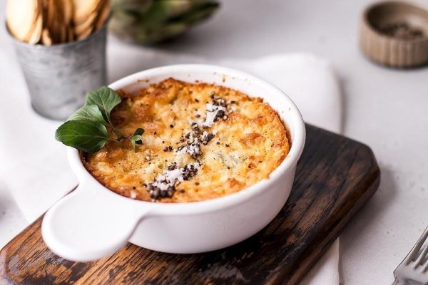 Artichoke dip in a white bowl on a wooden cutting board.