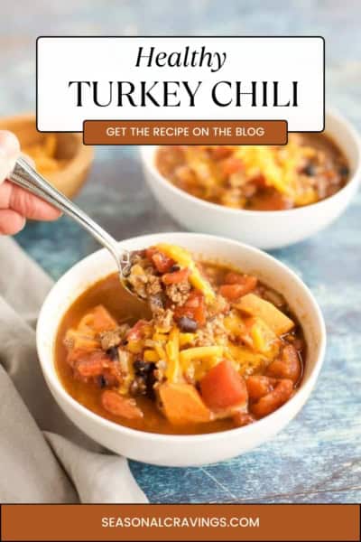 A hand holding a spoon over a bowl of healthy turkey chili topped with shredded cheese, with two other bowls of chili in the background. The text reads "Healthy Turkey Chili. Get the recipe on the blog.