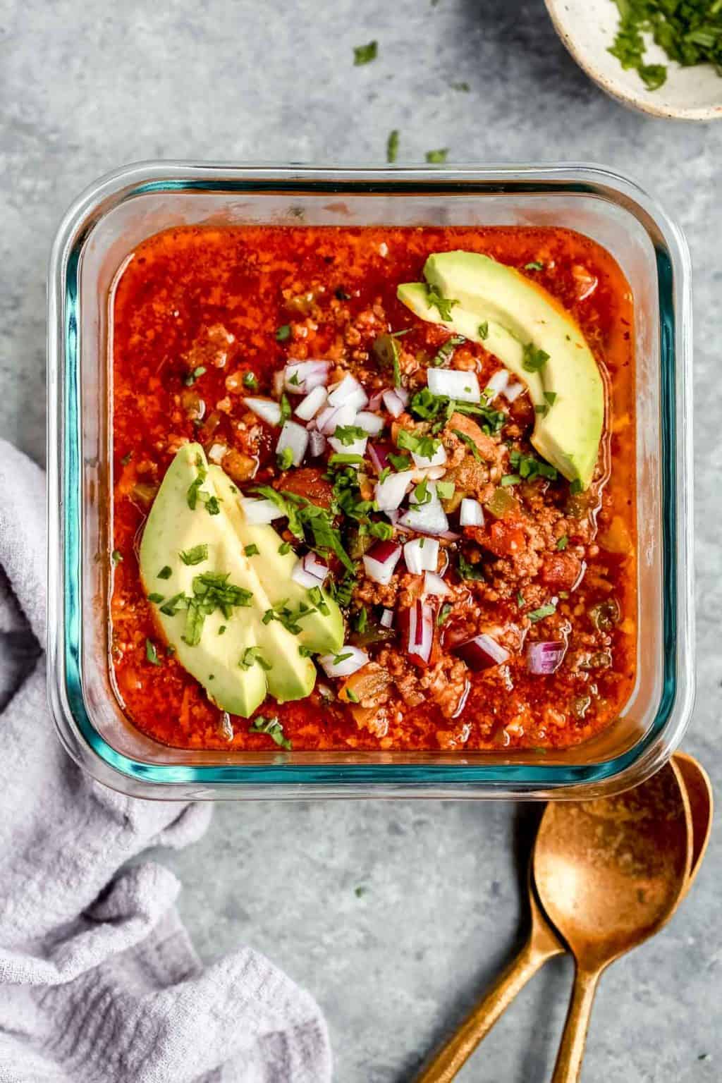 glass container holding one serving of chili with avocado slices and onions.
