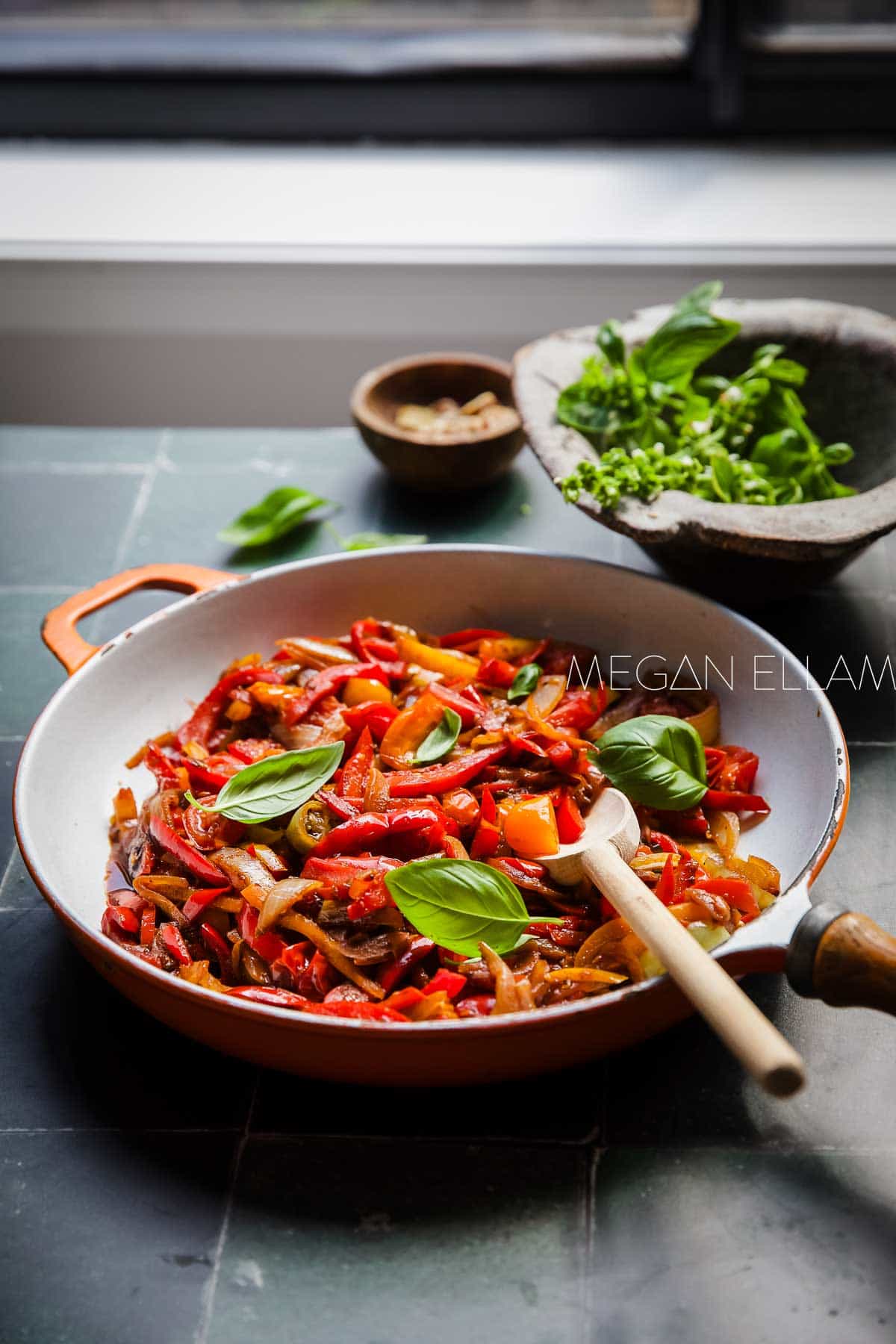 A bright orange skillet on a green tiles near a window filled with Italian pepper stew. 
