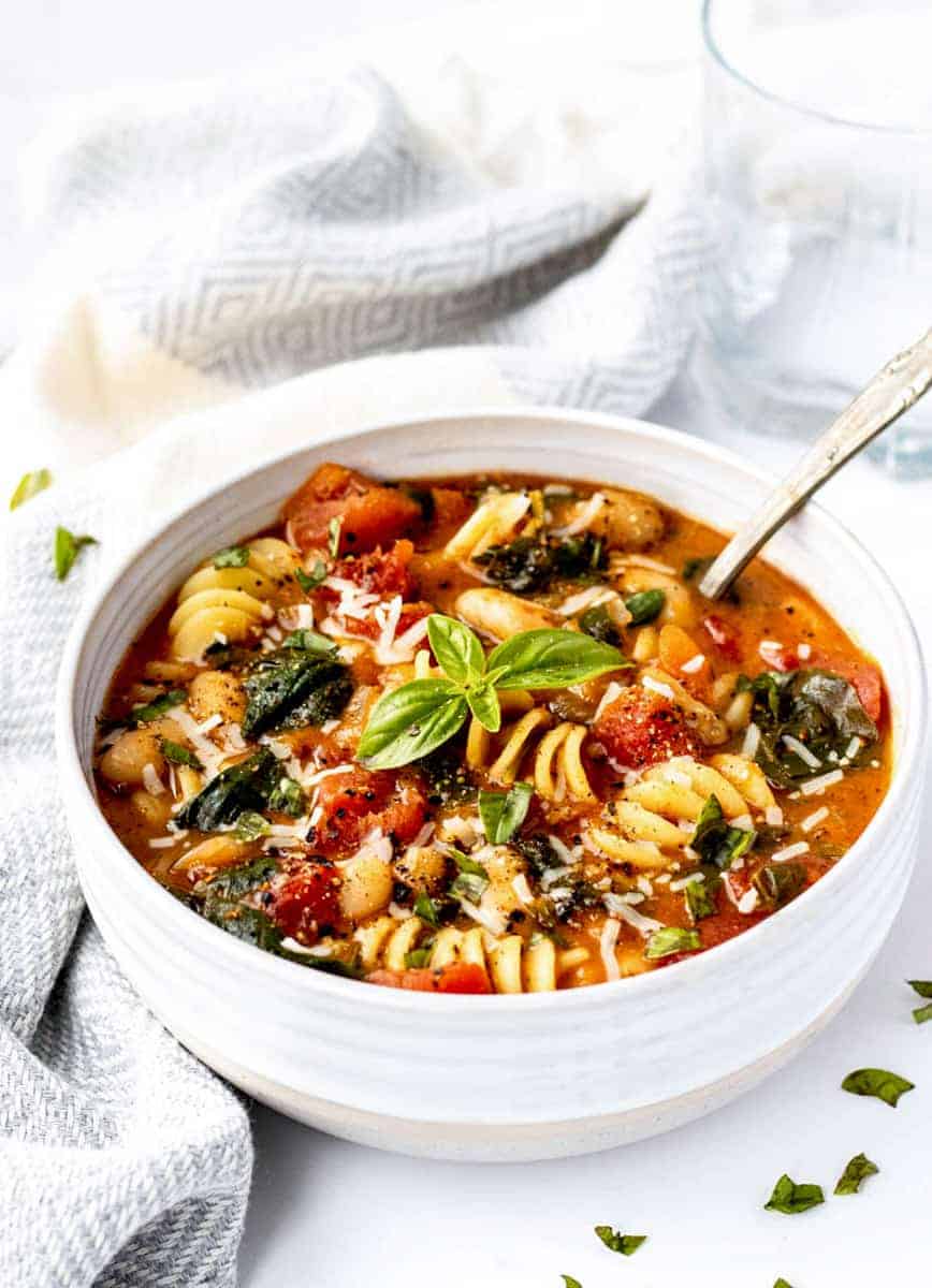 Tuscan white bean soup served in a white bowl with a spoon and fresh basil. 
