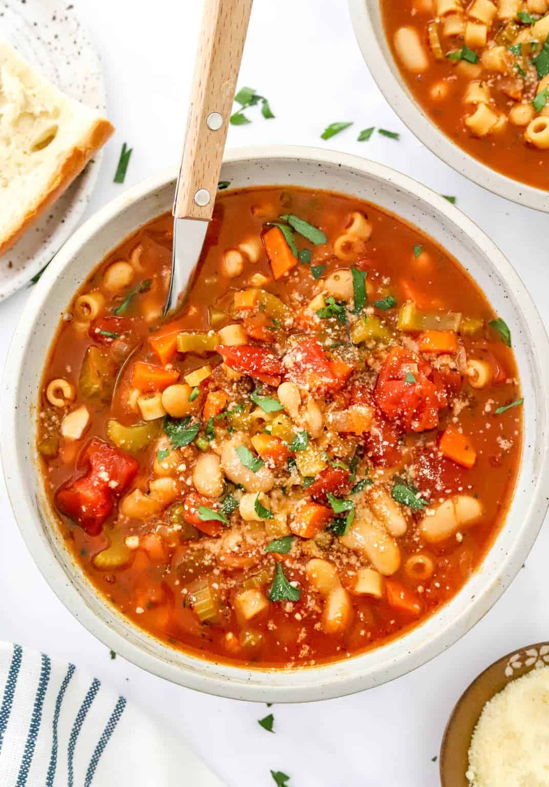 Vegetarian pasta fagioli in a bowl with spoon. Crusty bread, parmesan, and linen towel on a counter. 
