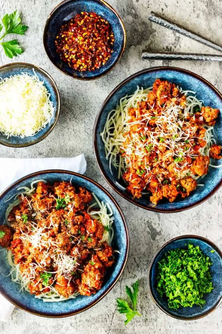 Overhead photo of two bowls of keto spaghetti surrounded by small bowls of parsley, shredded parmesan, and crushed red pepper flakes. 
