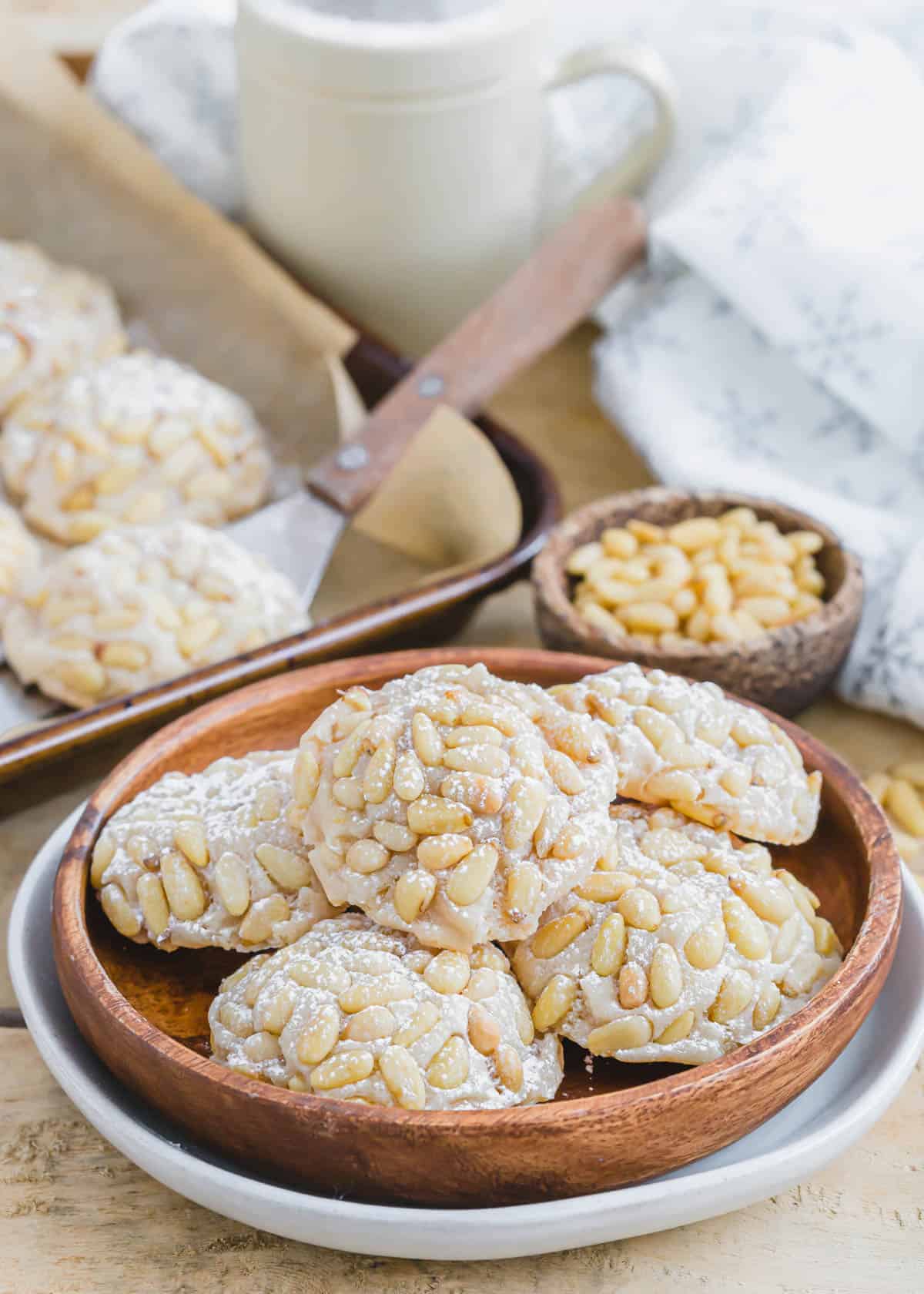 Vegan pignoli cookies on a wooden plate with pine nuts in the background. 
