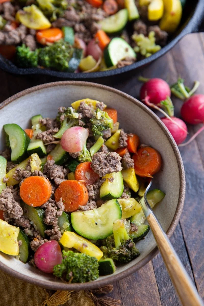 Bowl of beef and vegetables with a skillet of beef and vegetables in the background. 
