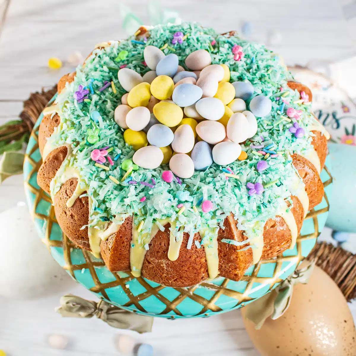 Easter mini egg cake topped with lemon icing, coconut, and Cadbury mini eggs on cake stand with easter decor. 
