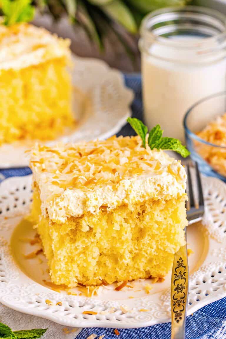 Side view of two pieces of pina colada poke cake served on plates with forms, surrounded by a bowl of toasted coconut and a glass of milk. 
