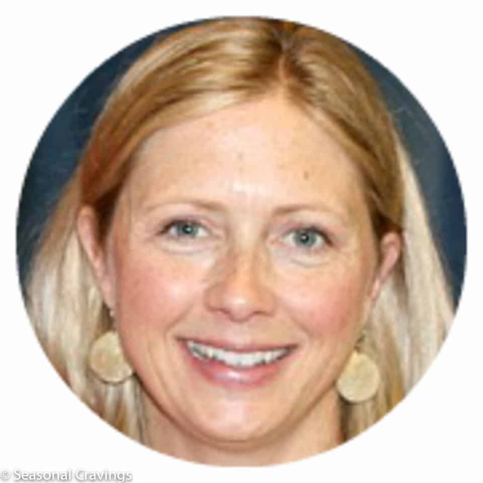 A woman with blonde hair smiling at the camera. She is wearing circular earrings and has a dark background behind her.